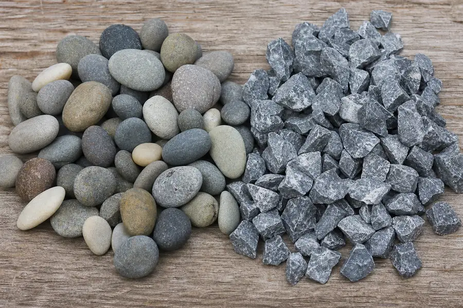 Side-by-side handful of rounded river gravel and angular crushed stone on a wooden surface